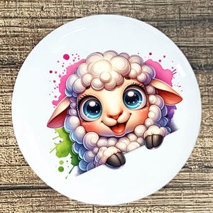 May include: White ceramic plate featuring a cartoon sheep with large blue eyes, a pink nose, and a smiling expression. The sheep is surrounded by pink and green watercolor splashes. The plate has a round shape.
