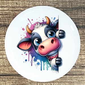 May include: A white ceramic plate featuring a cartoon cow peeking from behind a white surface. The cow has black and white markings, large blue eyes, and a pink nose. Colorful watercolor paint splatters in pink, purple, blue, and teal surround the cow.