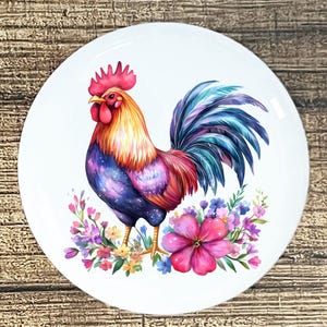 May include: A white ceramic plate featuring a colorful rooster illustration. The rooster has a red comb and wattle, with a vibrant mix of blue, purple, orange, and yellow feathers. The plate is decorated with flowers.
