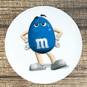 May include: A round white ceramic plate featuring a centered graphic of the blue M&M character. The character is oval-shaped, colored blue, and displays a white lowercase letter m on its front, with tan arms, white gloves, and gray shoes.