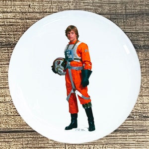 May include: A white round button with a picture of a character from Star Wars wearing an orange jumpsuit and holding a helmet. The character has brown hair and is standing on a white background.
