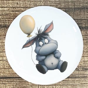 May include: White ceramic plate featuring a cartoon illustration of a blue donkey holding a yellow balloon. The donkey has a sad expression and is sitting down. The plate has a glossy finish.
