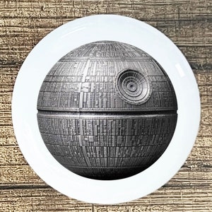 May include: A white ceramic plate featuring a detailed image of the Death Star from Star Wars. The Death Star is rendered in shades of gray and has intricate surface details. The plate is round and sits on a wooden surface.