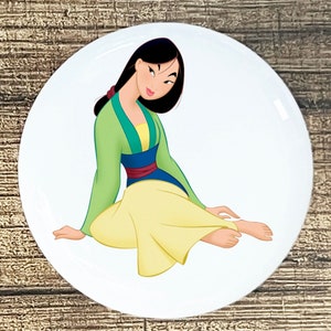 May include: A white button with a cartoon image of Mulan, a Disney princess, sitting with her legs crossed. She is wearing a green and yellow dress with a blue sash. Her hair is black and pulled back in a ponytail.