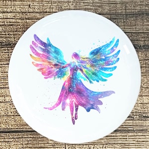 May include: White round decorative plate featuring a colorful watercolor image of an angel with outstretched wings. The angel's wings and dress are a blend of blues, purples, pinks, and yellows, with a galaxy-like design. The plate has a glossy finish.