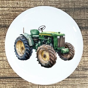 May include: A white ceramic plate featuring a watercolor illustration of a green tractor with black tires and yellow rims. The tractor is centered on the plate, showcasing its details against a plain white background. The plate has a glossy finish.