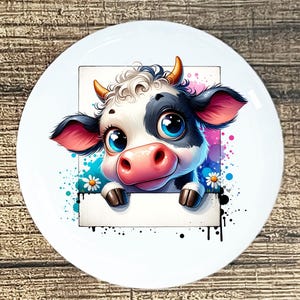 May include: A white ceramic plate featuring a cartoon cow illustration. The cow has a black and white pattern, large blue eyes, and a pink nose. The background includes colorful paint splatters and daisies.