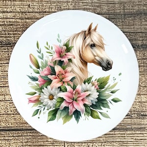 May include: A white ceramic plate featuring a horse's head surrounded by pink lilies, white daisies, and green foliage. The horse has a light brown coat and flowing white mane. The plate has a glossy finish.