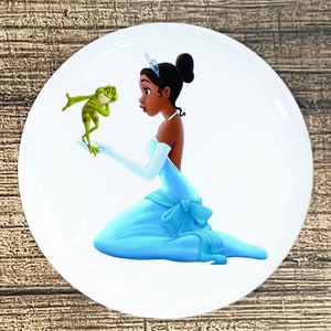 May include: A princess in a blue gown with a green frog standing in front of her. The princess has a crown on her head and is looking at the frog.
