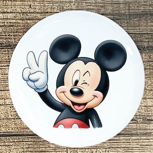 May include: White ceramic plate featuring a smiling Mickey Mouse character. Mickey is winking and giving a peace sign with his gloved hand. He is wearing a black jacket, red shorts, and has large black ears.