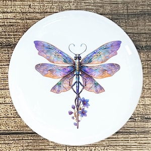 May include: White ceramic plate featuring a detailed illustration of a dragonfly. The dragonfly has intricate wings in shades of purple, blue, and orange. A sprig of blue flowers extends from the dragonfly's body. The plate is round and sits on a wooden surface.