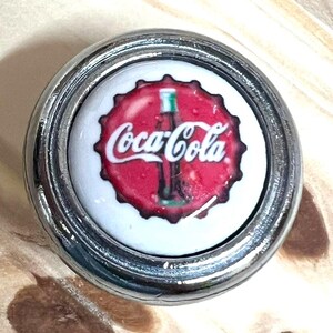 May include: A circular metal knob featuring a white center insert with a red bottle cap graphic. The graphic displays a dark glass bottle silhouette and the white Coca-Cola logo. The metallic frame has a polished silver finish with a raised edge.