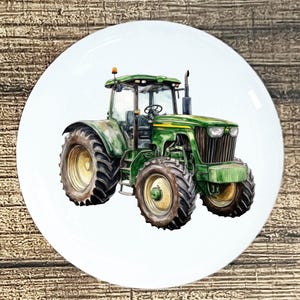 May include: A white ceramic plate featuring a watercolor illustration of a green tractor. The tractor has large tires, a cab, and is set against a white background. The plate is round and has a glossy finish.