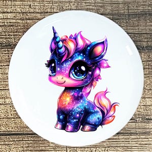 May include: A whimsical illustration of a cartoon unicorn with a galaxy-themed design. The unicorn features a blue horn, a pink and purple mane, and large, expressive eyes. The image is set against a white circular background.