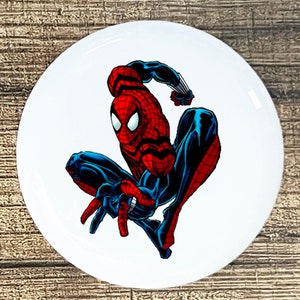 May include: A red and blue cartoon superhero, Spiderman, is in a crouching pose with his arms outstretched. He is wearing a red and blue suit with a black spider symbol on his chest.