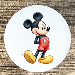 May include: A white, round plate featuring a smiling Mickey Mouse cartoon character. Mickey is depicted in his classic outfit: red shorts, yellow shoes, and black gloves. The plate has a glossy finish.