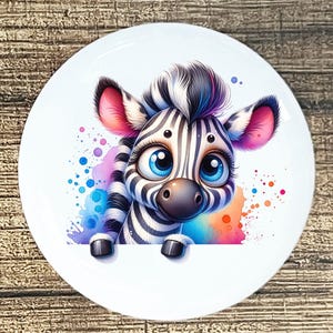 May include: A white ceramic plate featuring a cartoon zebra with large blue eyes and a black and white striped pattern. The zebra is set against a colorful watercolor splash of pink, blue, and orange.