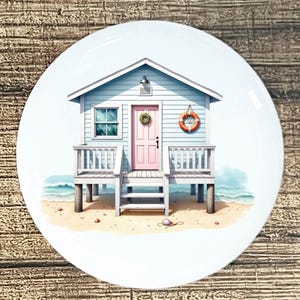 May include: A white ceramic plate featuring a beach scene. A light blue beach house with a pink door, white railings, and a life preserver is depicted on a sandy beach with ocean waves. A decorative wreath hangs on the door.