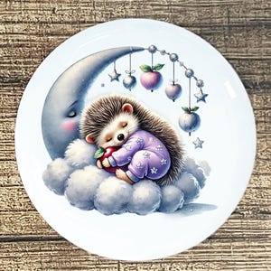 May include: A white ceramic plate featuring a whimsical illustration. A sleeping hedgehog in purple pajamas with star accents rests on a cloud, cradling an apple. A crescent moon and hanging stars and apples complete the design.