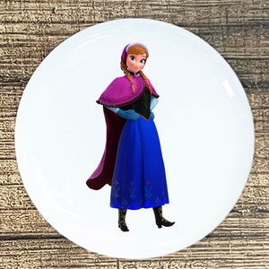 May include: A white button with a cartoon image of a girl with long brown hair wearing a purple and black dress and a blue cape. She is standing with her hands on her hips.