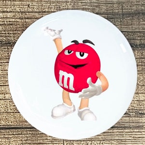 May include: A red M&M's character with a white smile and the letter "M" on its chest, waving with its right hand.