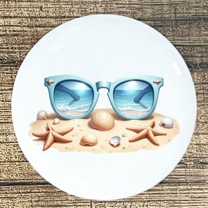 May include: White ceramic plate with a beach-themed design. Light blue sunglasses reflect a beach scene with waves. Starfish and seashells are scattered on a sandy beach. The plate has a glossy finish.