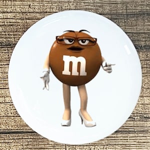 May include: A brown M&M character wearing white gloves and high heels. The character has a serious expression and is pointing to the right. The character has a white "M" on its chest.