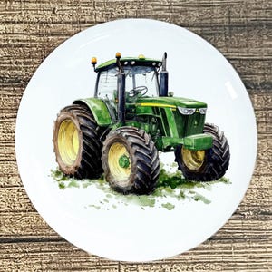 May include: A white ceramic plate featuring a watercolor illustration of a green tractor with black tires and yellow rims. The tractor is set against a backdrop of green grass and a wooden surface.