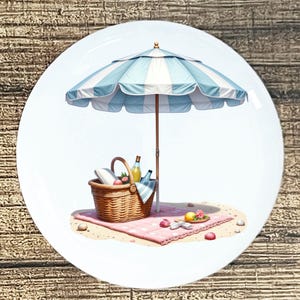 May include: A round white plate featuring a beach picnic scene. A blue and white striped umbrella shades a picnic basket, blanket, and food items. The scene is set on a sandy beach with seashells and a pink blanket.