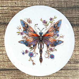 May include: A white, round decorative plate featuring a detailed butterfly design. The butterfly has orange, blue, and purple wings with intricate patterns. Delicate purple and blue flowers and green leaves surround the butterfly.