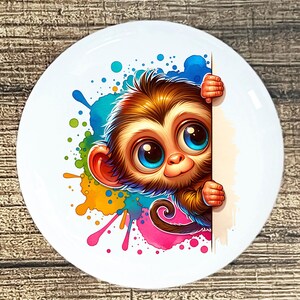 May include: White ceramic plate featuring a cartoon monkey with big blue eyes and brown fur. The monkey peeks from behind a white surface, surrounded by colorful paint splatters in blue, green, yellow, and pink.