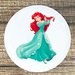May include: A cartoon illustration of Ariel, the Little Mermaid, wearing a teal gown with a white and silver pattern. She has long red hair and is holding a brush in her hand.