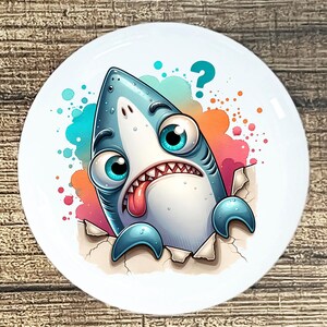 May include: A white ceramic plate featuring a cartoon shark bursting through a torn surface. The shark is blue and white with large, expressive eyes, a lolling tongue, and a question mark above. Colorful paint splatters surround the shark.