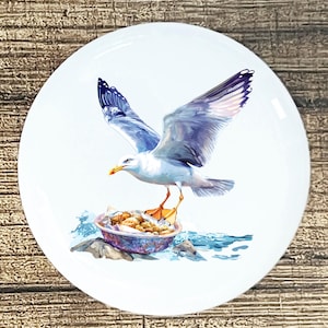 May include: White ceramic plate featuring a detailed illustration of a seagull in flight. The seagull has white and blue feathers, with a yellow beak and orange feet. The plate also depicts a bowl of food and ocean waves.