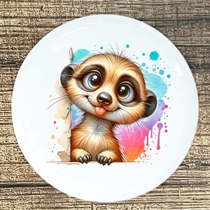 May include: A white ceramic plate featuring a cartoon meerkat with large blue eyes and a pink tongue. The meerkat is brown and tan with black ears and paws. Colorful watercolor splashes of blue, pink, and purple decorate the background.
