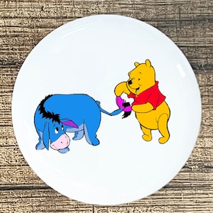 May include: A cartoon image of Winnie the Pooh pulling on the tail of Eeyore, a donkey. Winnie the Pooh is yellow and wearing a red shirt. Eeyore is blue and has a sad expression.