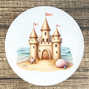 May include: A white ceramic plate featuring a whimsical illustration of a sandcastle on a beach. The castle has tan walls, brown conical roofs, and pink flags. A starfish and seashell are on the sand, with blue ocean waves in the background.