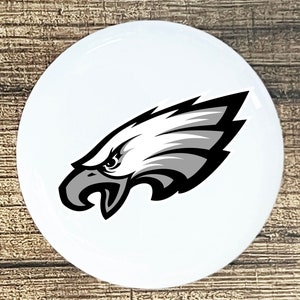 May include: A white round button with a black and gray eagle head logo. The eagle is facing right and has a sharp beak and fierce eyes.