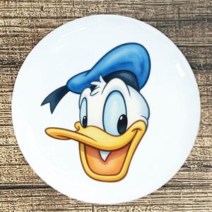 May include: White ceramic plate featuring a cartoon illustration of Donald Duck's face. The character has a blue sailor hat, a yellow beak, and a smiling expression. The plate is round and sits on a wooden surface.