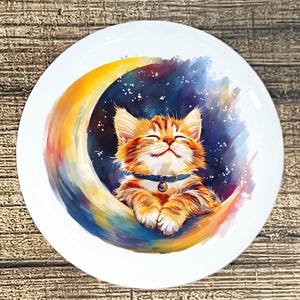 May include: A white ceramic plate featuring a whimsical illustration of a ginger tabby cat smiling serenely while nestled in a colorful crescent moon. The moon is painted with shades of yellow, orange, blue, and purple, set against a starry night sky.