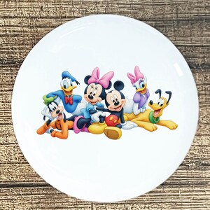 May include: White ceramic plate featuring a colorful illustration of Disney characters, including Mickey Mouse, Minnie Mouse, Donald Duck, Daisy Duck, Goofy, and Pluto. The plate has a glossy finish and is suitable for serving food.