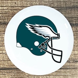 May include: A white ceramic plate featuring the Philadelphia Eagles football team logo. The logo depicts a teal football helmet with white wings and a black facemask. The plate has a glossy finish.