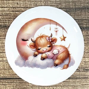 May include: A white ceramic plate featuring a whimsical illustration of a sleeping Highland cow calf. The calf, in a pink onesie, rests on a cloud against a crescent moon. Decorative elements include a star and bows.