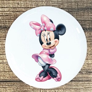 May include: A white ceramic plate featuring a smiling Minnie Mouse illustration. Minnie Mouse is depicted in a pink dress and bow with white polka dots. The plate has a glossy finish and is suitable for serving food.