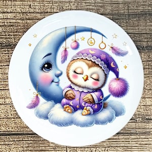 May include: A round white plate featuring a whimsical illustration. A sleepy owl in purple pajamas and nightcap rests on a cloud next to a smiling blue moon. Decorative elements include hanging feathers, stars, and pom-poms.