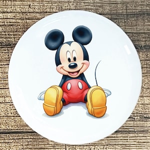 May include: A white, round plate featuring a smiling Mickey Mouse illustration. Mickey is depicted in his classic attire: red shorts, yellow shoes, and black ears. The plate is set against a wooden background.