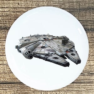 May include: A white round button with a gray and white spaceship, the Millennium Falcon, from Star Wars.