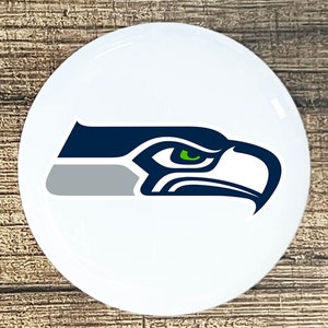 May include: A white round button with a blue and green Seattle Seahawks logo. The logo is a stylized image of a hawk's head with a green eye.