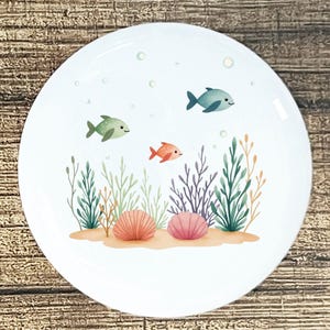 May include: White ceramic plate with an underwater scene. The design features three cartoon fish in shades of green, orange, and blue, swimming above colorful seaweed and two seashells. Bubbles float near the top of the plate.