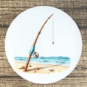 May include: A white ceramic plate featuring a beach scene with a fishing rod. The rod is brown with a blue handle and a pink bobber. The scene includes a sandy beach, seashells, and blue ocean waves.